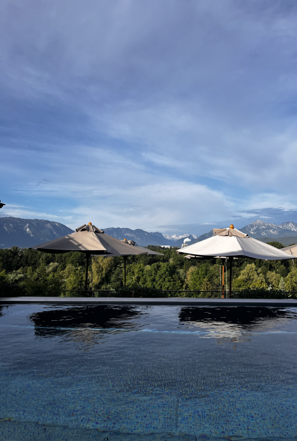 Pool mit Sonnenschirmen, dahinter Wald und Berge unter blauem Himmel