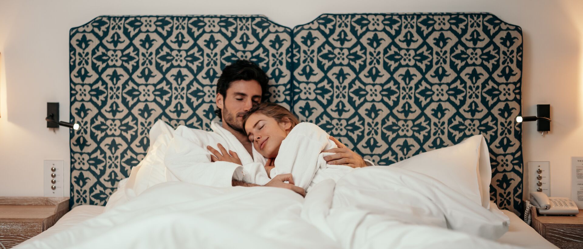 Paar in weißen Bademänteln kuschelt im Bett mit gemustertem Kopfteil