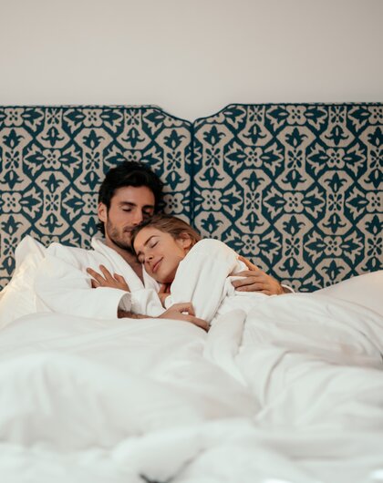Paar in weißen Bademänteln kuschelt im Bett mit gemustertem Kopfteil