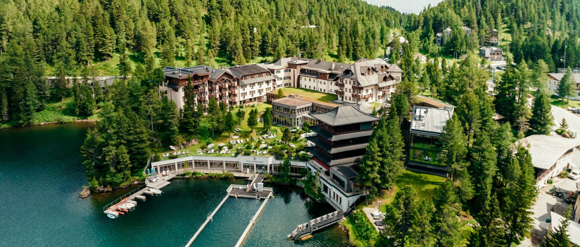 A luxurious hotel resort is idyllically located by a clear mountain lake, surrounded by forests and mountains.