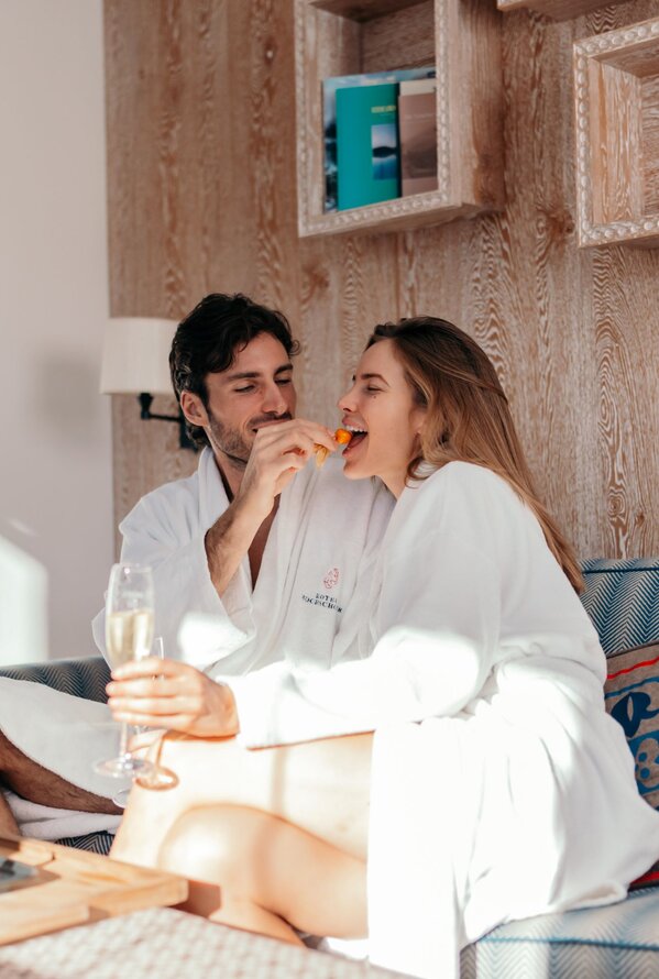 Ein Paar im Bademantel entspannt auf einem Sofa und genießt Obst und ein Getränk.