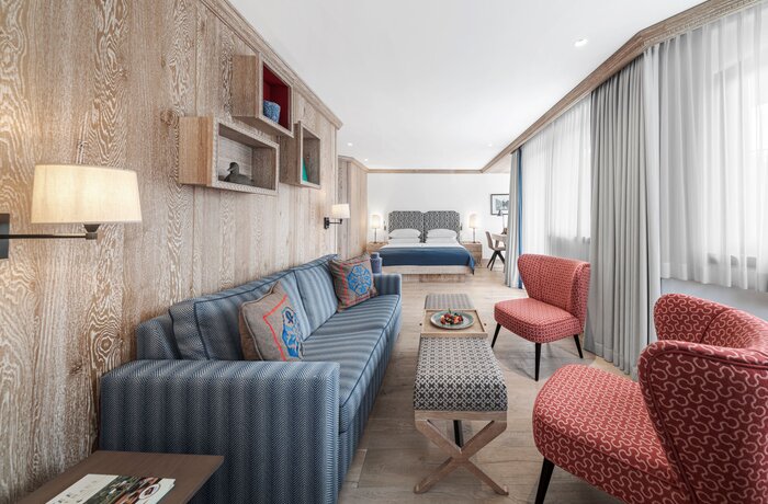 A modern hotel room with a sofa, armchairs, and a bed in the background. Large windows let in plenty of natural light.