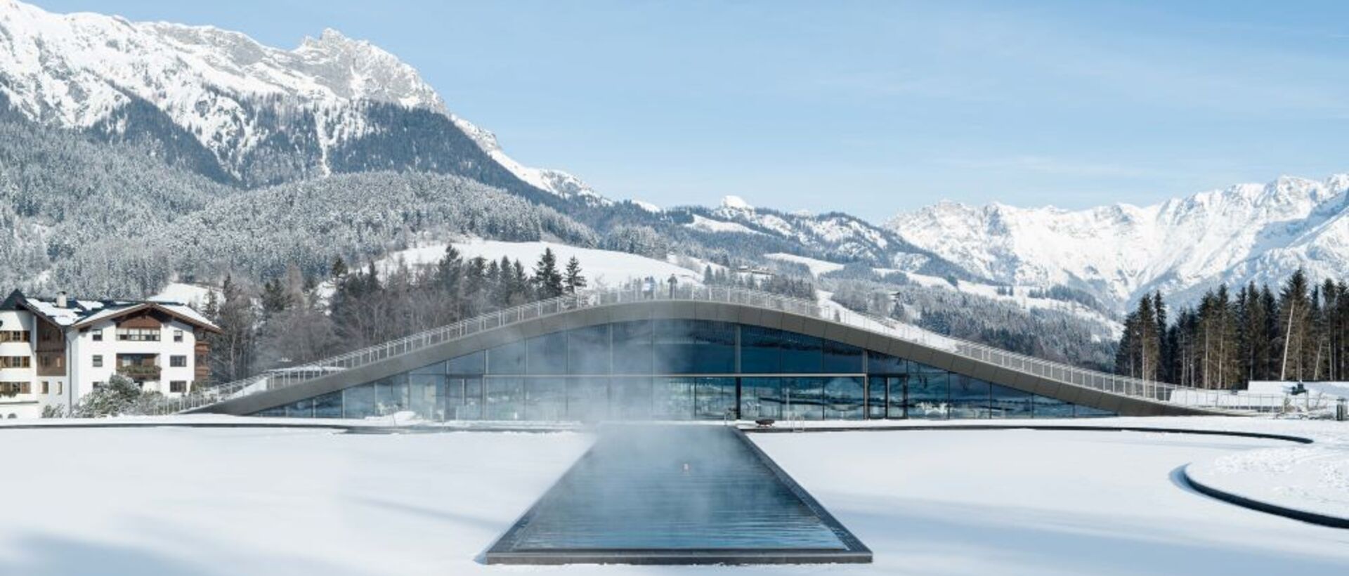 Winterlandschaft mit schneebedecktem Infinity-Pool vor Bergpanorama und modernem Gebäude