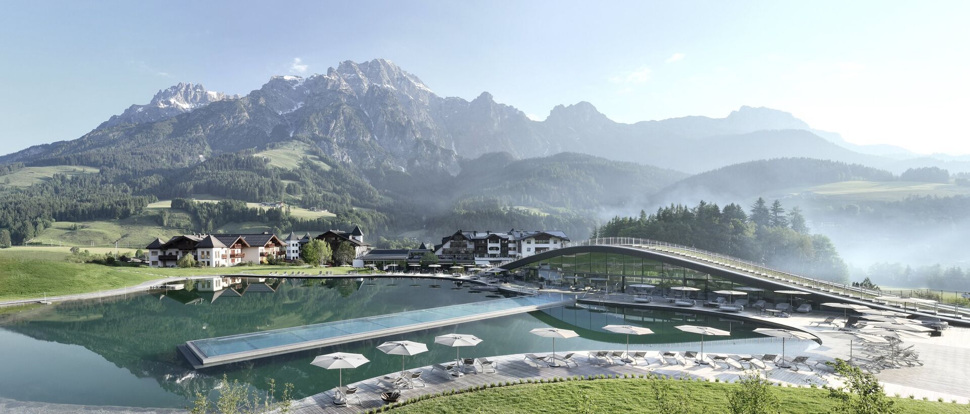 Badesee mit Liegen vor Bergpanorama und Gebäuden unter blauem Himmel