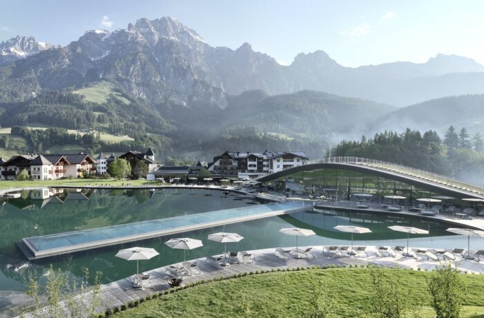 Badesee mit Liegen vor Bergpanorama und Gebäuden unter blauem Himmel