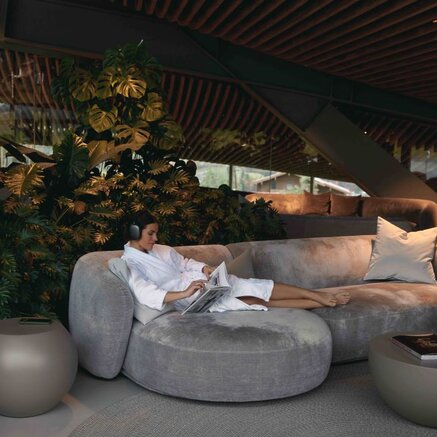 Woman in bathrobe reading on grey sofa with headphones in modern interior with plants