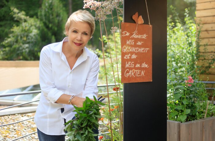 Frau mit kurzem blonden Haar im Garten, Schild mit Spruch: Der kürzeste Weg zur Gesundheit ist der Weg in den Garten
