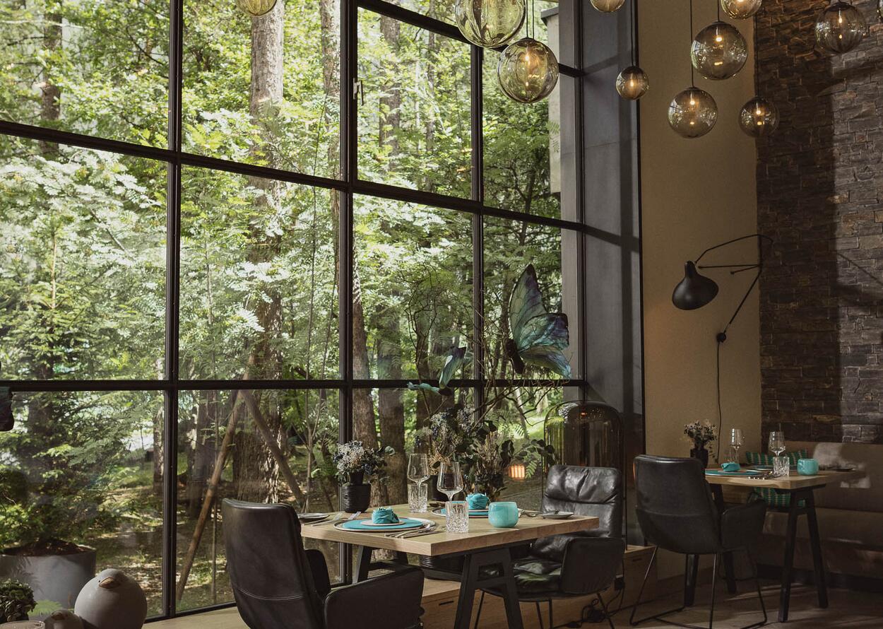 Modernes Restaurant mit großen Fenstern, warmem Licht und Blick in den Wald.