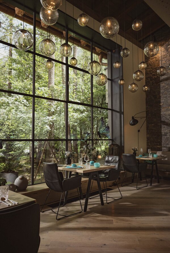 Modernes Restaurant mit großen Fenstern, warmem Licht und Blick in den Wald.