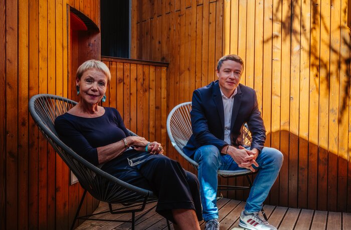 Eine ältere Frau und ein jüngerer Mann sitzen entspannt auf Stühlen in einem Innenhof mit hohen Holzwänden. Warmes Sonnenlicht und Schatten verleihen der Szene eine ruhige, stilvolle Atmosphäre.