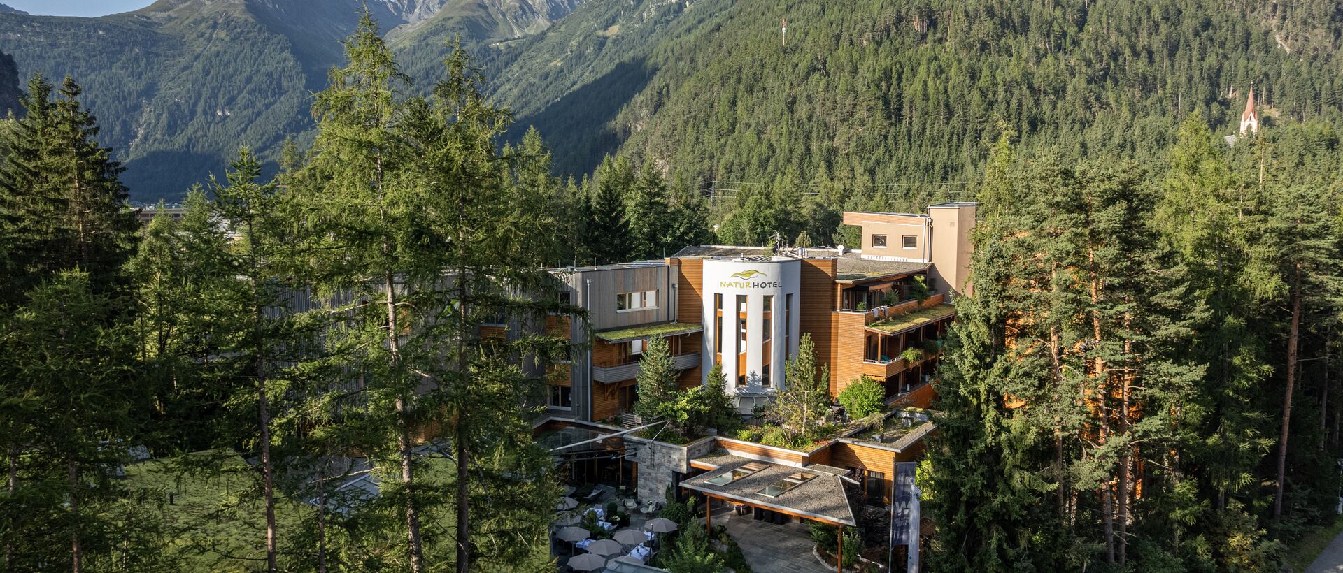 Ein modernes Hotel liegt eingebettet zwischen hohen Bäumen vor einer beeindruckenden Berglandschaft.