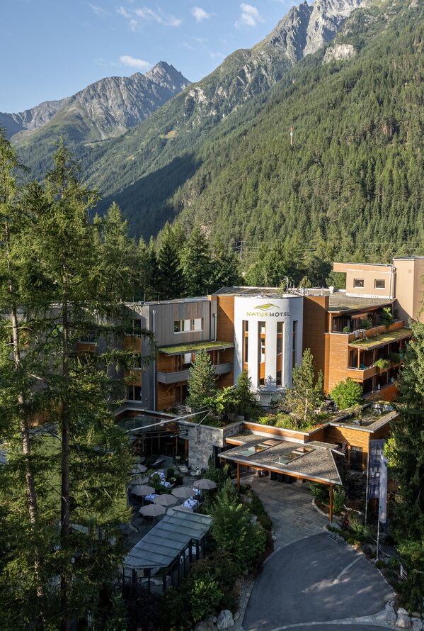 Ein modernes Hotel liegt eingebettet zwischen hohen Bäumen vor einer beeindruckenden Berglandschaft.
