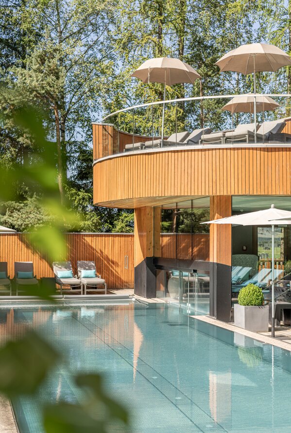 Ein ruhiger Außenpool liegt vor einem modernen Spa-Gebäude mit Holzfassade, Sonnenschirmen und Liegen auf einer Dachterrasse.