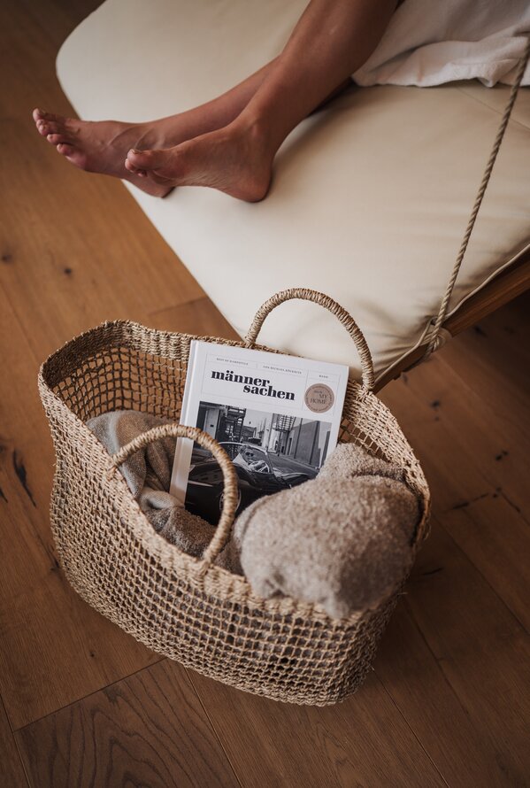 Eine Person entspannt auf einer Liege neben einem Korb mit Decke und Magazin.