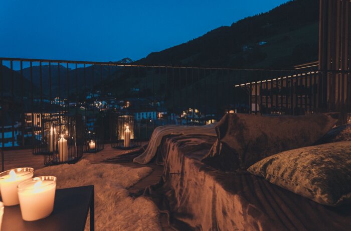A cozy terrace at night with candlelight, soft cushions, and a view over a lit mountain village. The warm lighting creates a relaxing and romantic atmosphere.