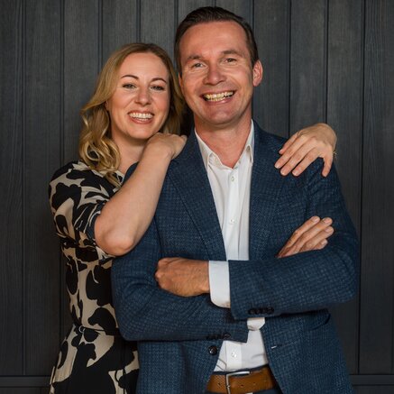 A smiling couple poses together in front of a dark wooden wall, with the woman resting her arms on the man's shoulders.
