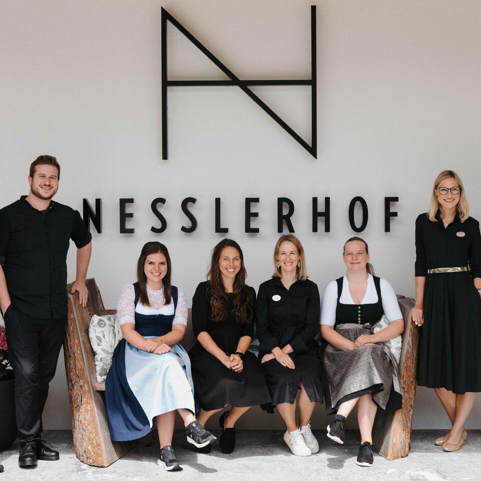 Freundliches Hotelteam posiert gemeinsam vor dem Nesslerhof-Logo.