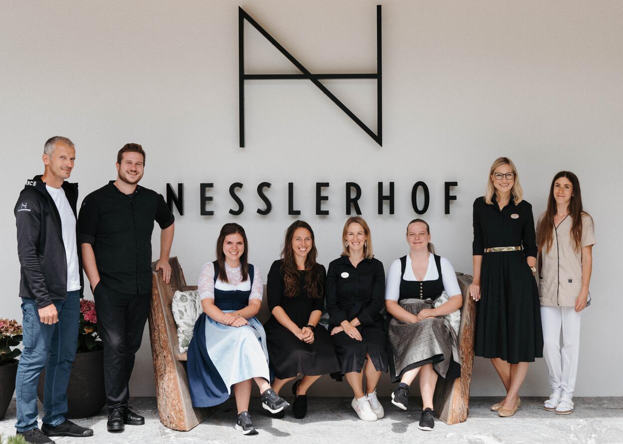 Freundliches Hotelteam posiert gemeinsam vor dem Nesslerhof-Logo.