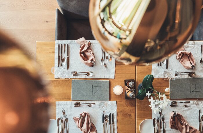 Gedeckter Holztisch mit Besteck, Servietten, Gläsern, Menükarten und Dekoration unter einer Lampe