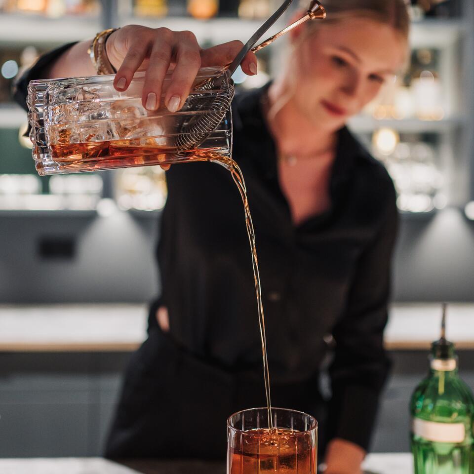 Eine Frau hinter einer Bar gießt einen bernsteinfarbenen Cocktail aus einem Glasgefäß in ein Trinkglas. Im Hintergrund stehen beleuchtete Flaschenregale.