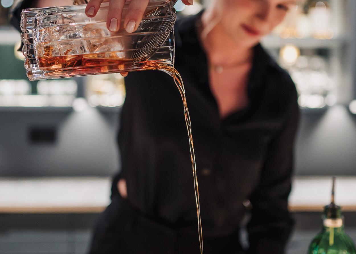 Eine Frau hinter einer Bar gießt einen bernsteinfarbenen Cocktail aus einem Glasgefäß in ein Trinkglas. Im Hintergrund stehen beleuchtete Flaschenregale.
