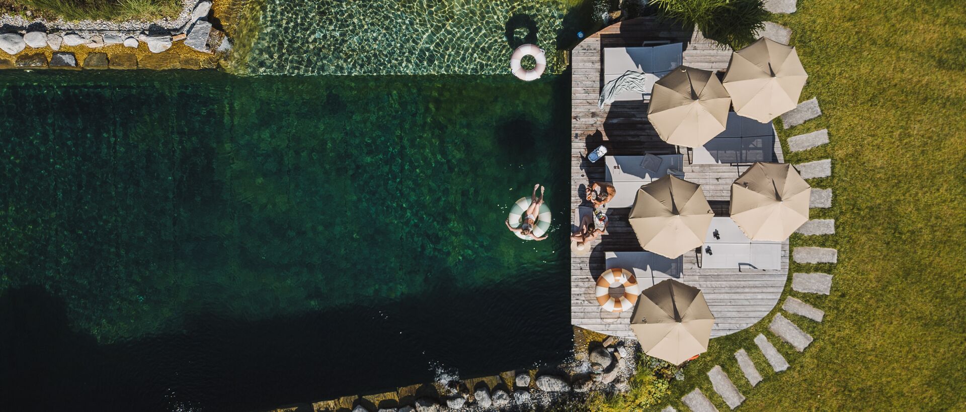 Drohnenaufnahme eines Naturpools mit Sonnenschirmen, Holzdeck und angelegtem Garten