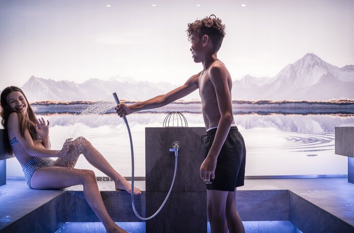 Junge duscht Füße einer Person in einem Innenpool vor einer Bergkulisse