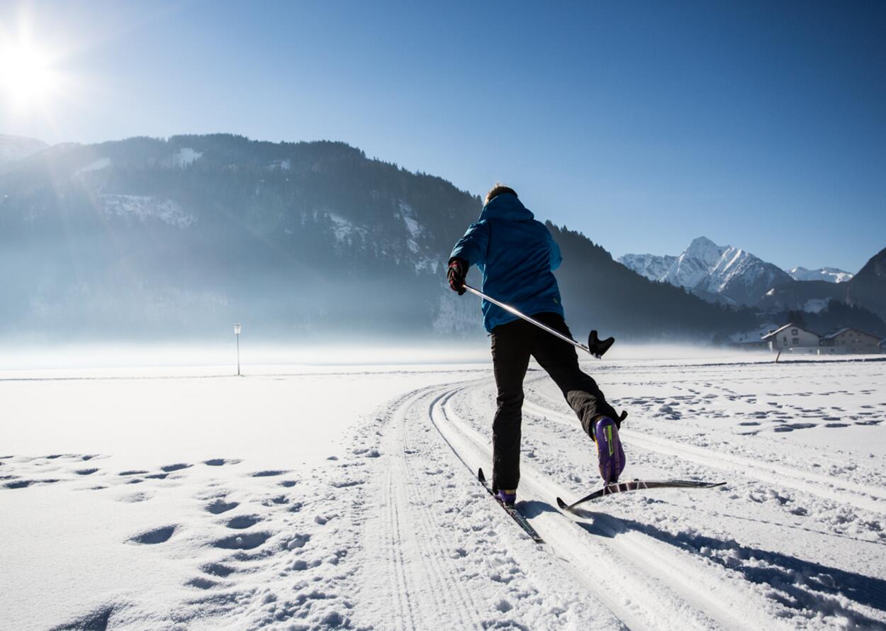 Eine Person fährt auf Langlaufskiern durch eine weite verschneite Landschaft, umgeben von Bergen und klarem Winterhimmel.