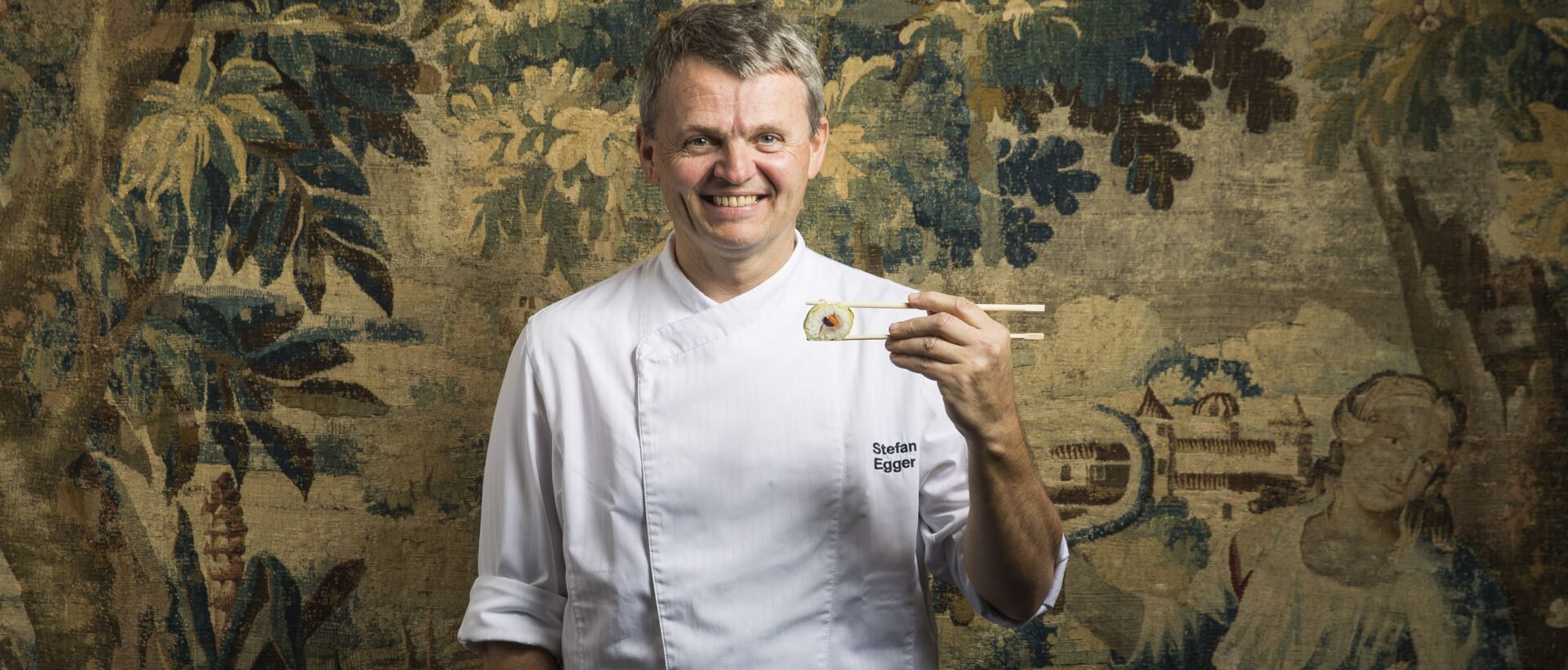 Chef Stefan Egger in white coat holding sushi with chopsticks in front of floral background