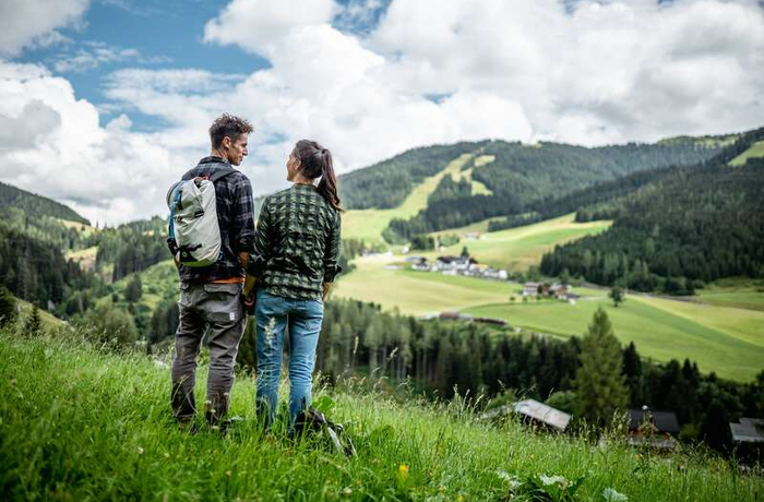 Hiking couple looking at green alpine meadow with houses and wooded hills under cloudy sky