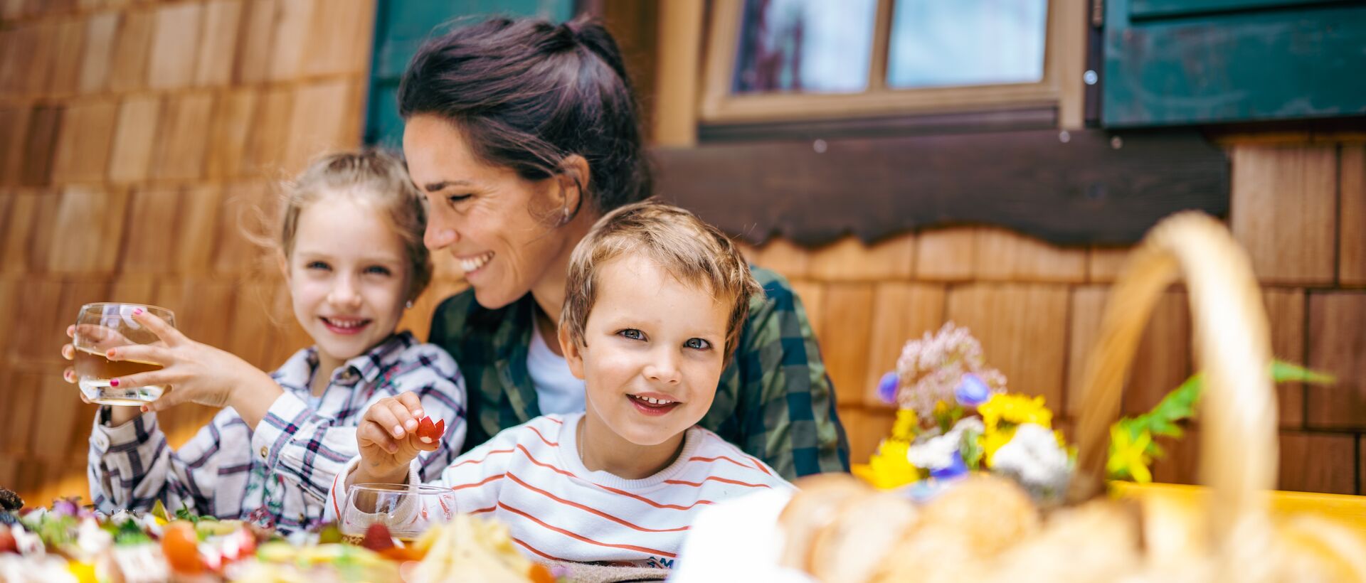 Frau und zwei Kinder sitzen lächelnd an einem gedeckten Tisch vor einem Holzhaus