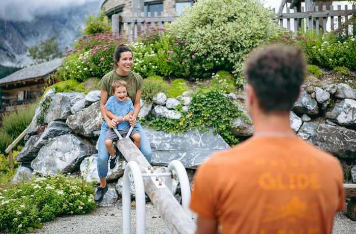 Eine Frau sitzt mit einem kleinen Kind auf einer Wippe aus Holz in einem naturnahen Spielbereich mit Steinen, Blumen und viel Grün. Im Vordergrund steht unscharf eine weitere Person, während im Hintergrund Gebäude und Berge zu sehen sind.