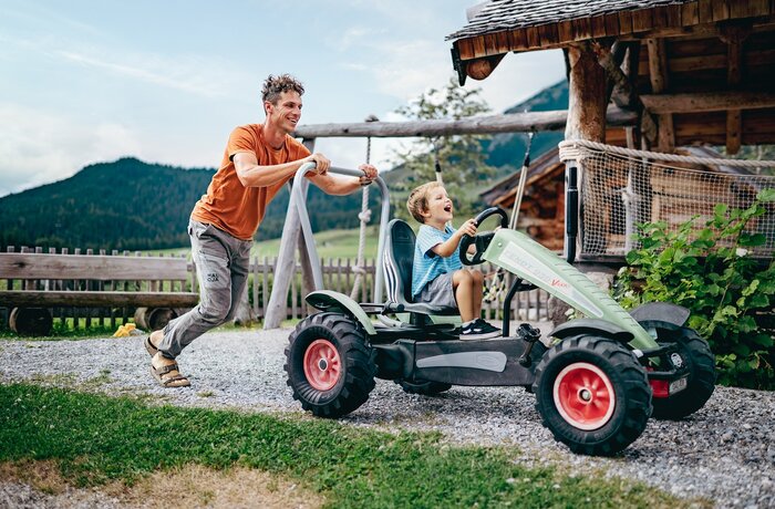Man pushes laughing boy in go-kart in front of wooden hut and mountain backdrop