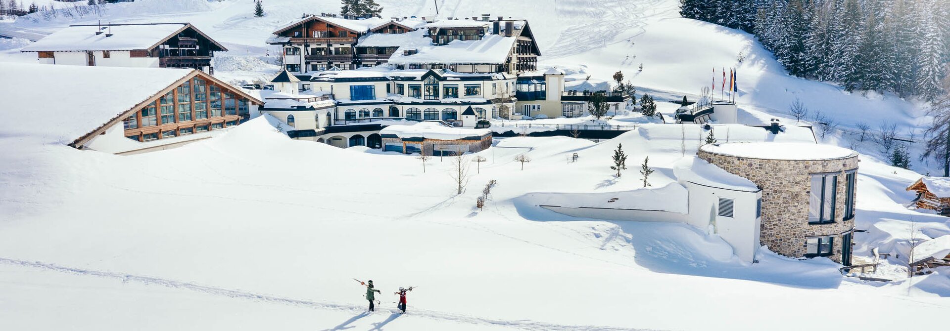 Hotel im Schnee mit Skipiste im Hintergrund und zwei Skifahrer im Vordergrund