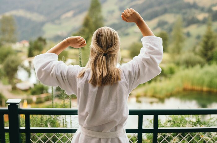 A woman in a white bathrobe stretches on a balcony overlooking mountains, nature, and water.