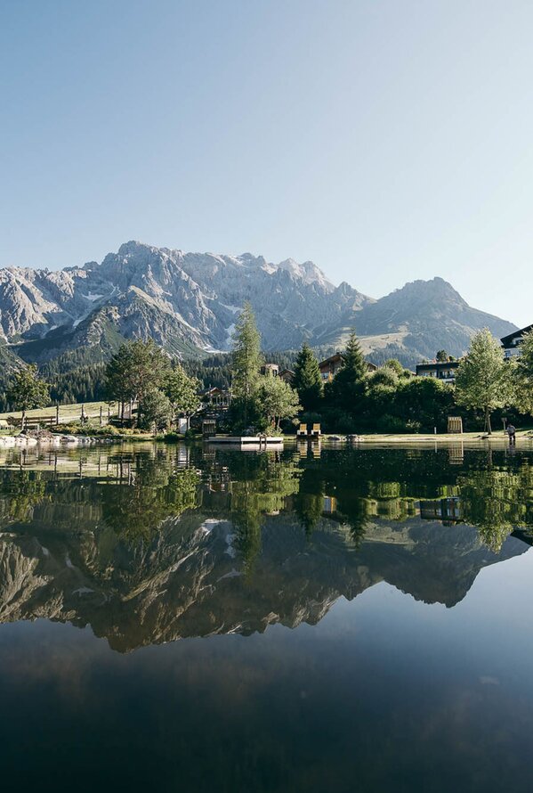 Ruhiger Bergsee mit Spiegelung der Alpen und umliegenden Bäume im Wasser.