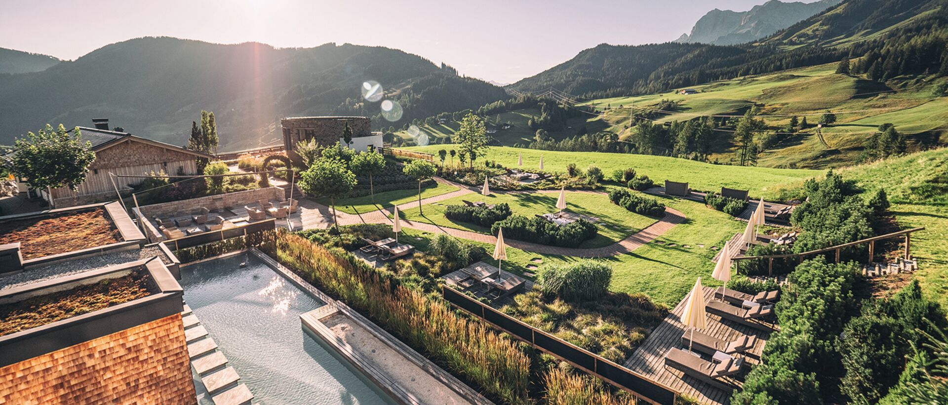 Ein langes Außenbecken mit Holzterrassen und Liegen liegt in einer weitläufigen Gartenlandschaft vor grünen Hügeln und Bergen im warmen Sonnenlicht.