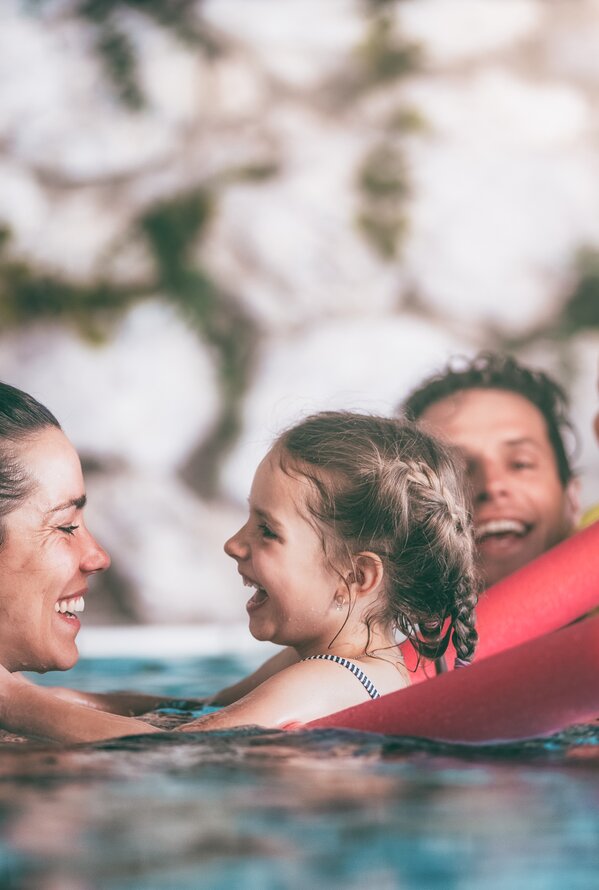 Eine Familie spielt und lacht gemeinsam im Wasser eines Pools mit bunten Schwimmnudeln.