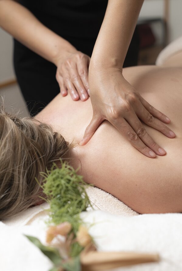 Eine Person liegt entspannt auf einer Massageliege, während ein Masseur mit beiden Händen den Rücken behandelt. Im Vordergrund liegen Kräuter und ein Handtuch.