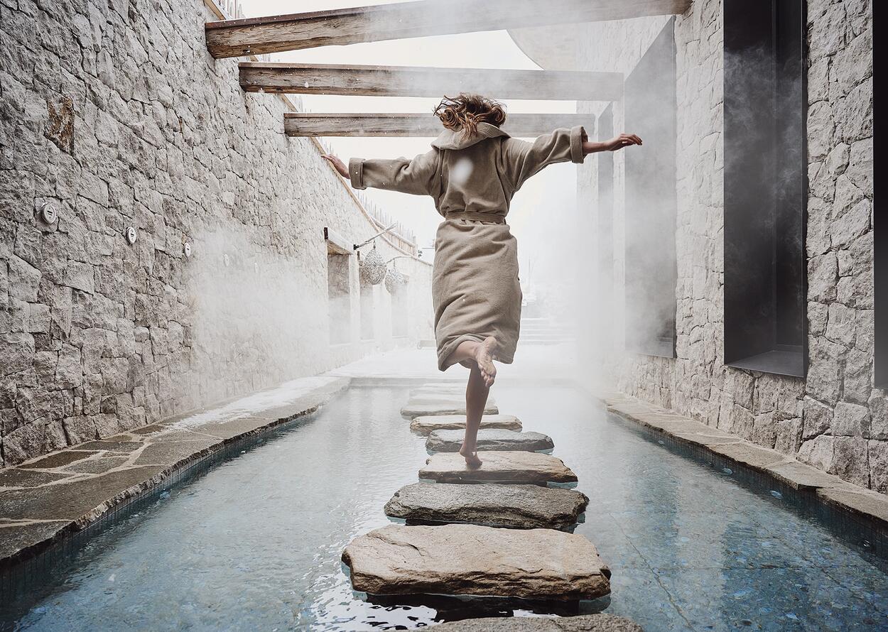 Woman in bathrobe balancing barefoot on stones over water in spa