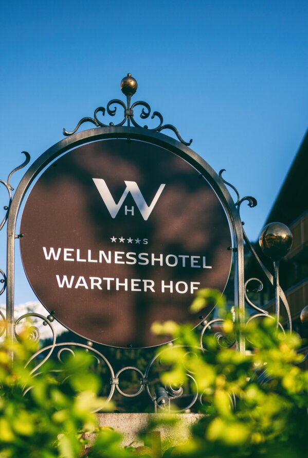 Das Eingangsschild des Wellnesshotels Warther Hof vor dem Hotelgebäude an einem sonnigen Tag.