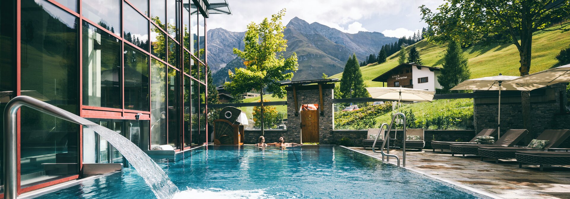Schwimmbad vor Bergkulisse mit Glasfassade und Wasserfall im Warther Hof