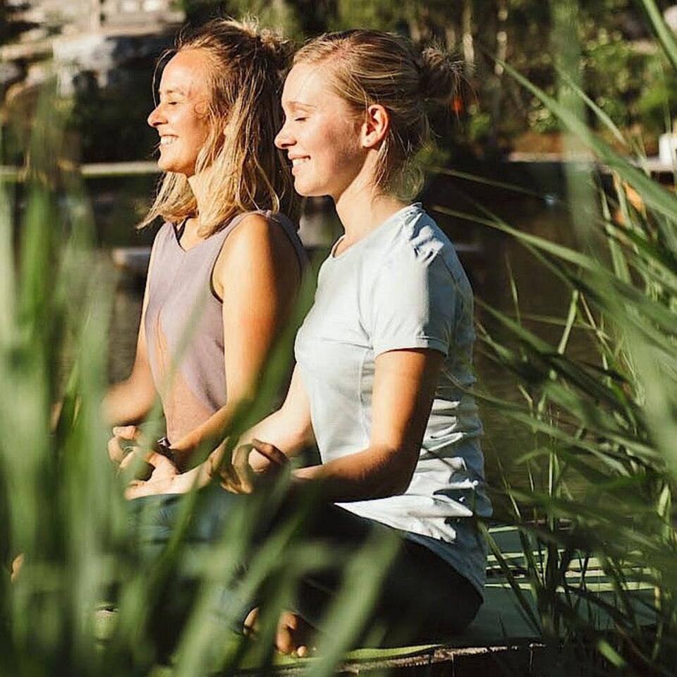 Zwei Frauen meditieren lächelnd im Freien vor Schilfgras
