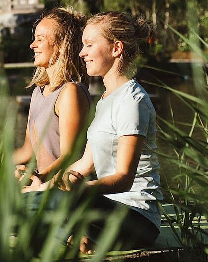 Zwei Frauen meditieren lächelnd im Freien vor Schilfgras