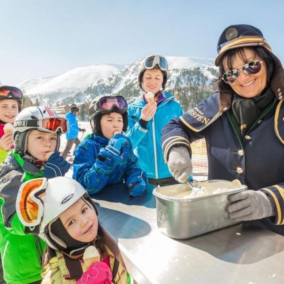 Frau verkauft Eis an Kinder in Skikleidung vor Bergkulisse