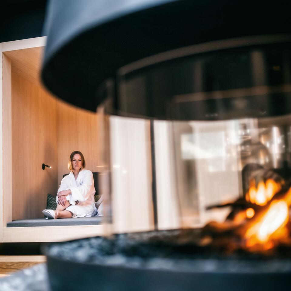 Frau im Bademantel sitzt in Nische, Kaminfeuer im Vordergrund