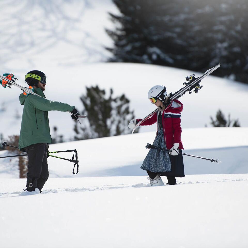 Two people in the snow, one with skis on his shoulder, the other with ski poles