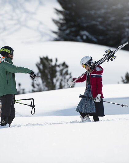 Two people in the snow, one with skis on his shoulder, the other with ski poles