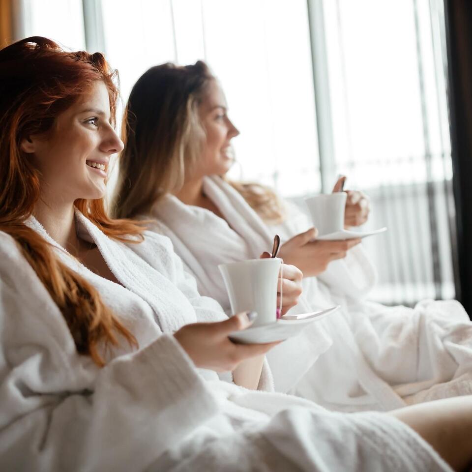 Two women in bathrobes with cups sitting relaxed in front of a window