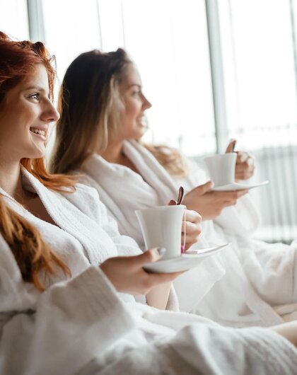 Two women in bathrobes with cups sitting relaxed in front of a window
